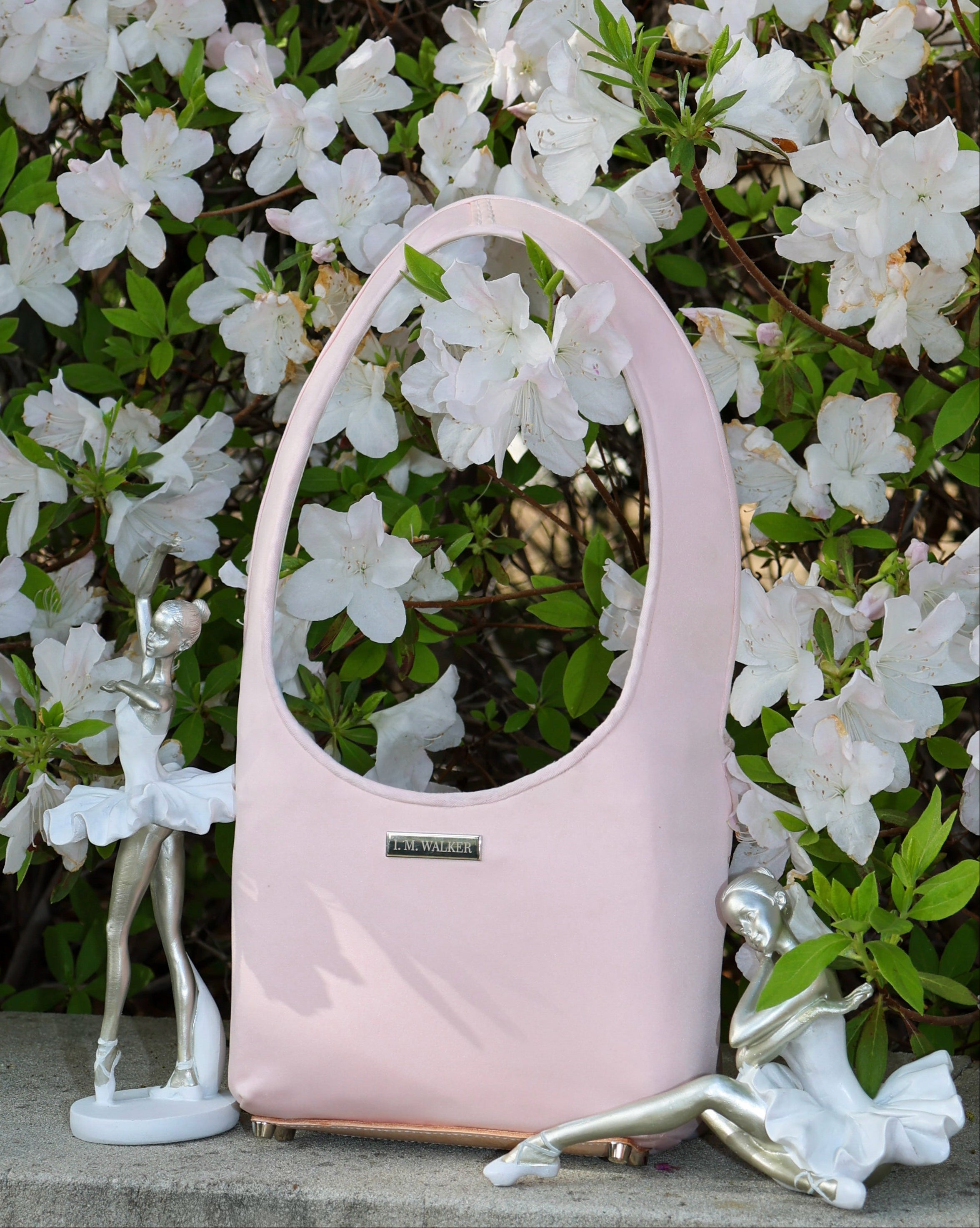 Pink handbag on a stone surface with white flowers and greenery in the background and ballet figurine statues.